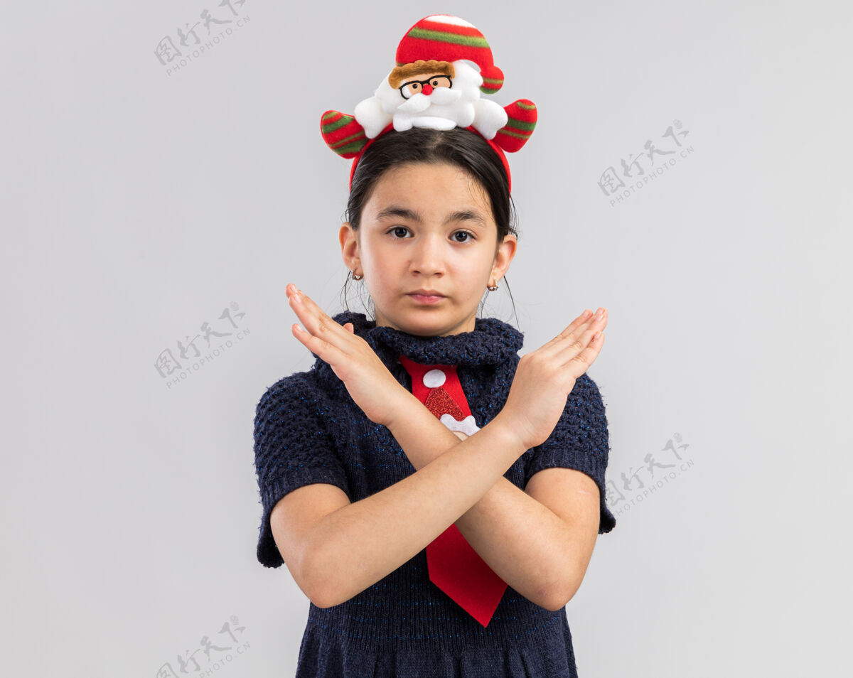 女孩小女孩穿着针织连衣裙 头上系着一条红色领带 头上戴着一个有趣的圣诞戒指 表情严肃 双手交叉 做着停下来的手势穿着交叉脸