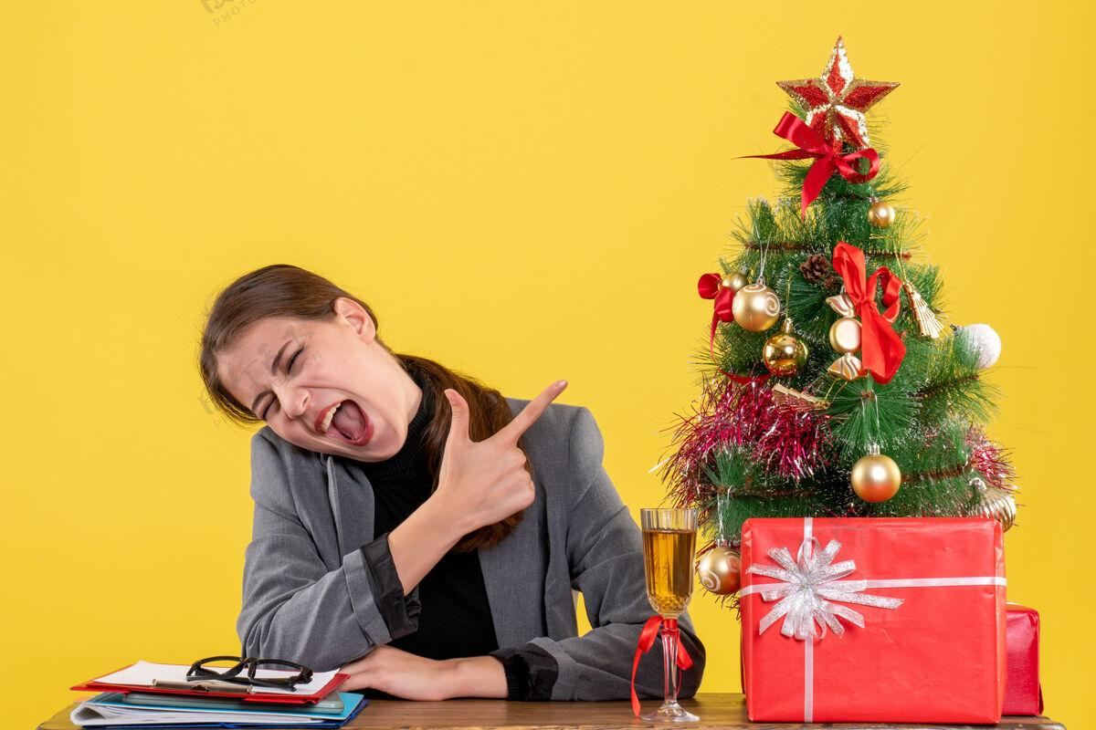 喊叫坐在书桌旁的女孩用手指着圣诞树和鸡尾酒礼物喊道手指表演者桌子