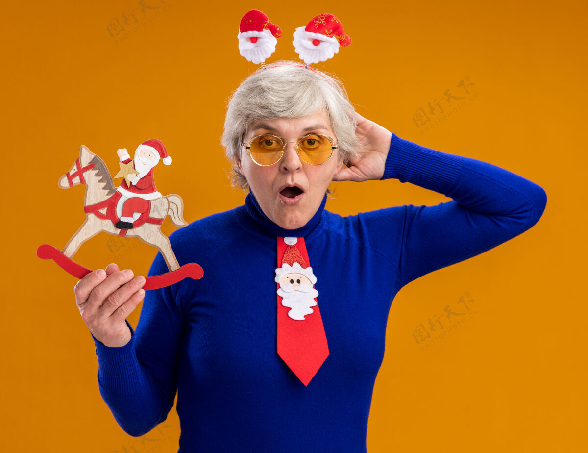 老人兴奋的老太婆戴着太阳镜 戴着圣诞老人的头带 打着圣诞老人的领带 把圣诞老人抱在摇摇木马上 装饰隔离在橙色的背景上 还有复制空间领带橙色抱着