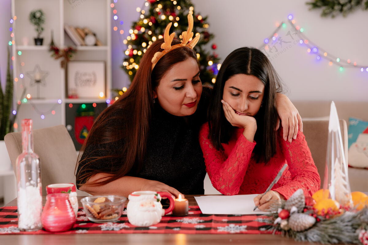看着高兴的母亲戴着驯鹿的头带看着女儿正在写的信坐在桌旁享受着家里的圣诞节时光桌子头带家