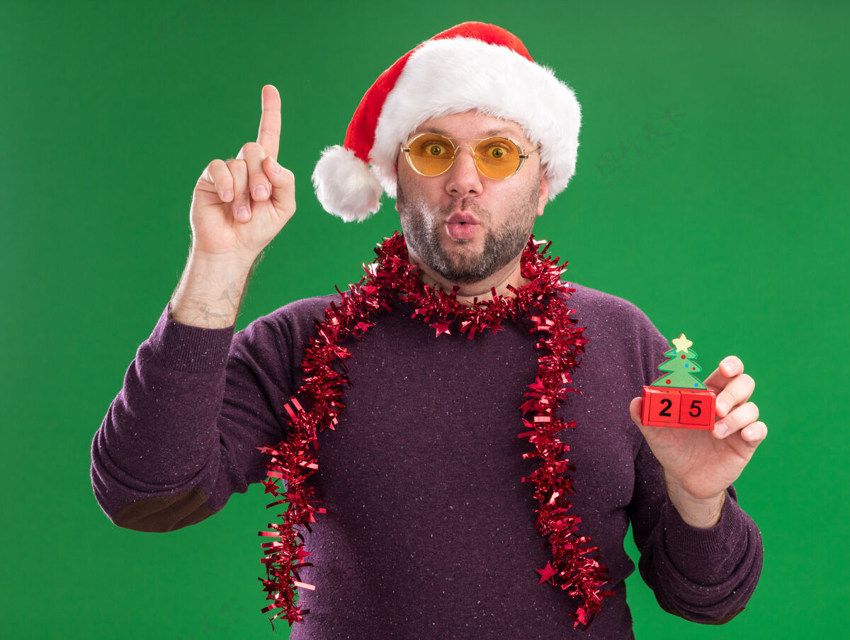 花环给人印象深刻的中年男子戴着圣诞帽 脖子上戴着金箔花环 戴着眼镜 手里拿着圣诞树玩具 日期朝上 噘着嘴唇 隔离在绿色的墙上帽子金属片中年人