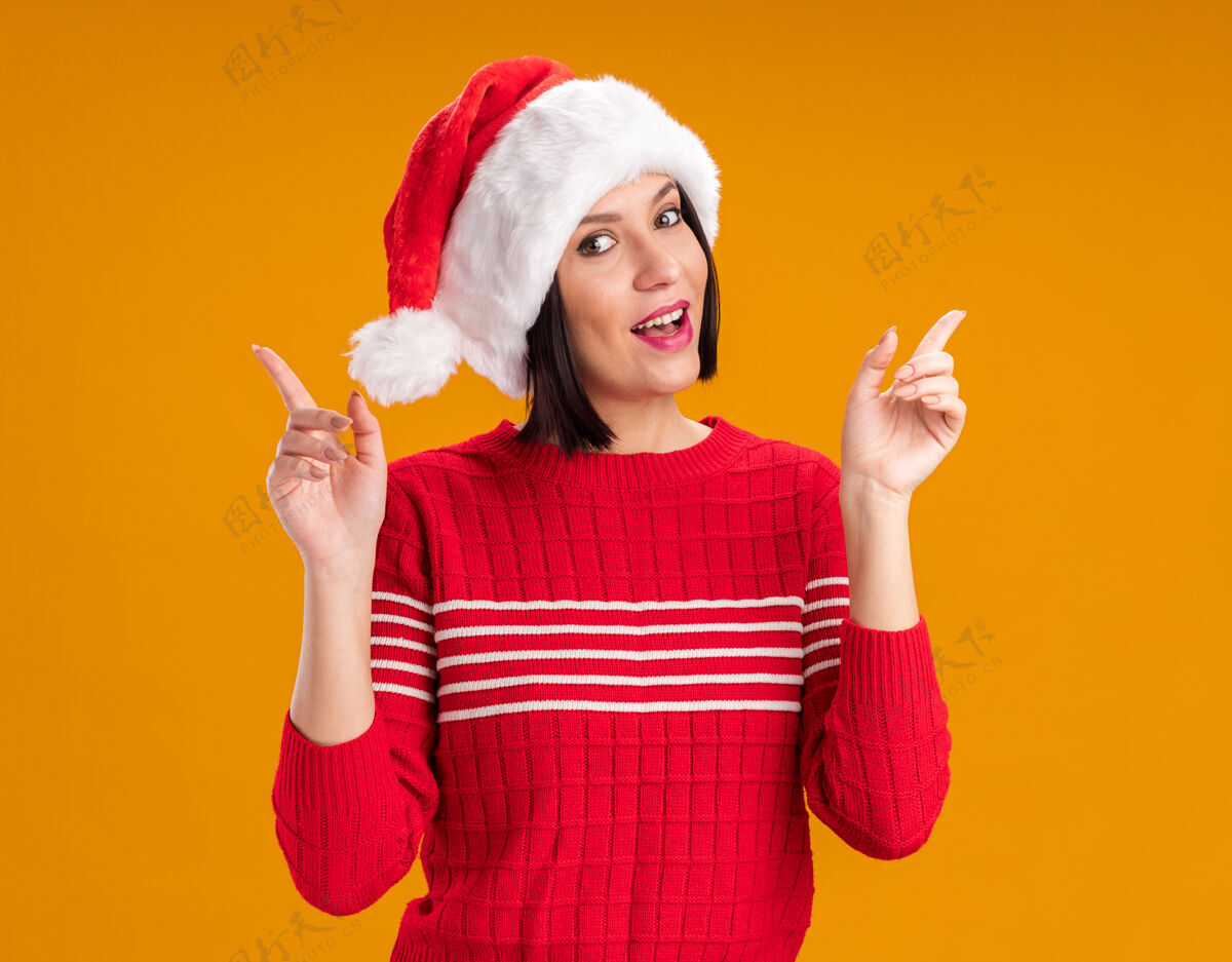 圣诞老人令人印象深刻的年轻女孩戴着圣诞帽指着橙色的墙上孤立年轻圣诞节戴着