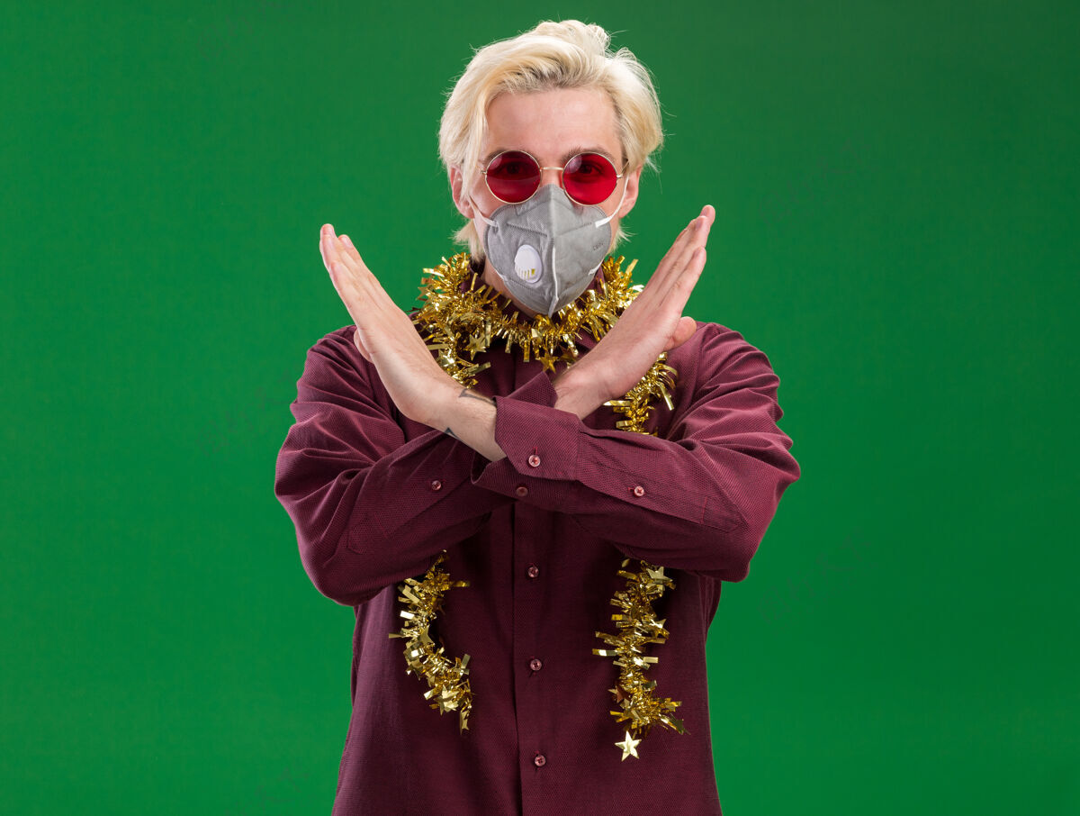 保护严肃的年轻金发男子戴着眼镜 戴着防护面具 脖子上戴着金属丝花环 在绿色的墙上做着不做任何动作周围圣诞节金属丝