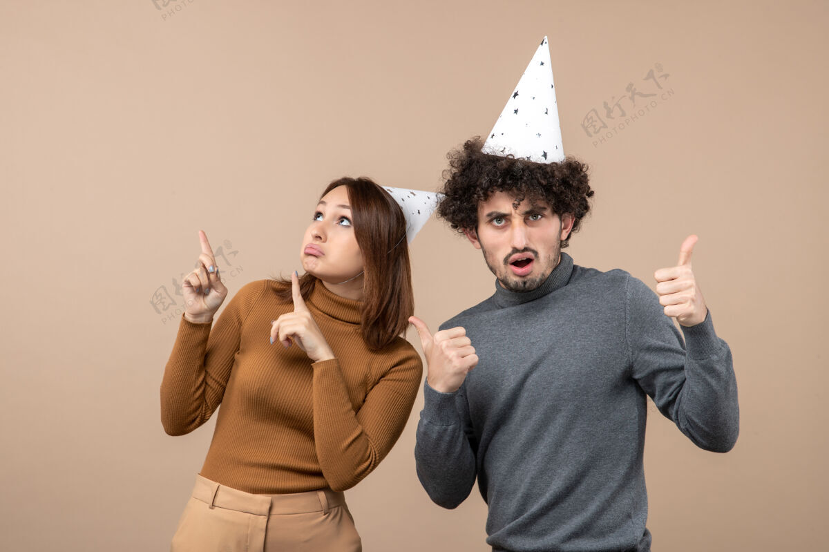 大人可爱兴奋的年轻夫妇戴着新年帽的女孩看上面和站在附近的灰色形象的家伙肖像帽子小伙子