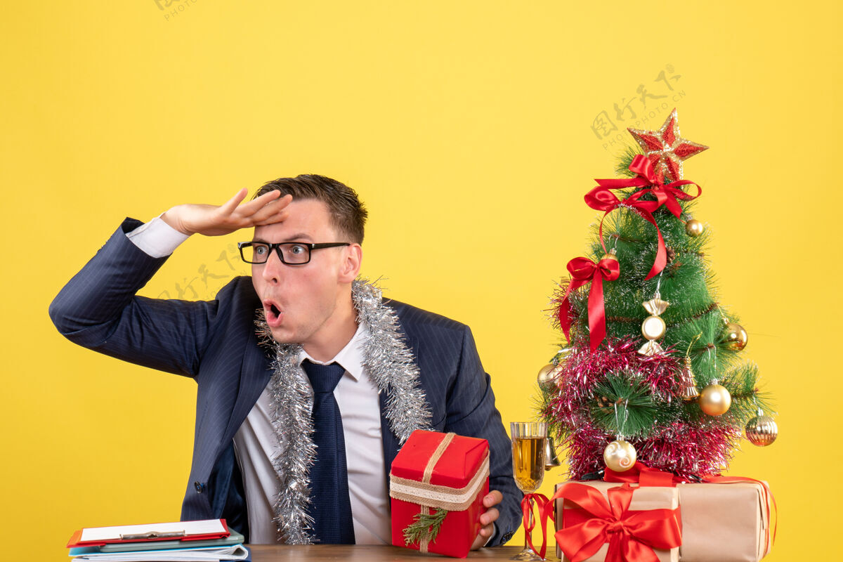 男前视图观察年轻人戴着眼镜坐在圣诞树旁的桌子上 黄色的礼物桌子树圣诞节