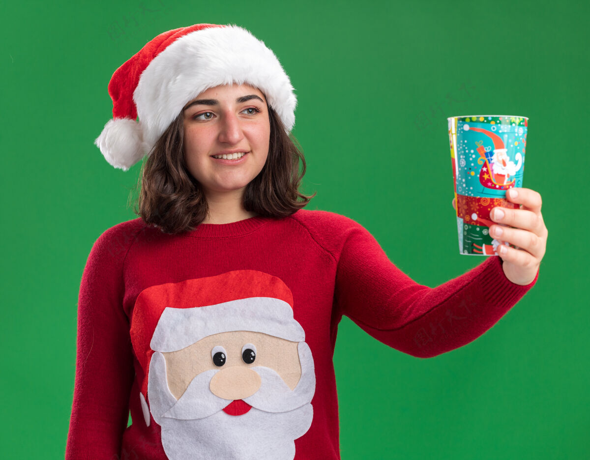 穿着快乐的小女孩穿着圣诞毛衣 戴着圣诞帽 手里拿着五颜六色的纸杯 面带微笑地看着它站在绿色的墙上年轻圣诞老人毛衣