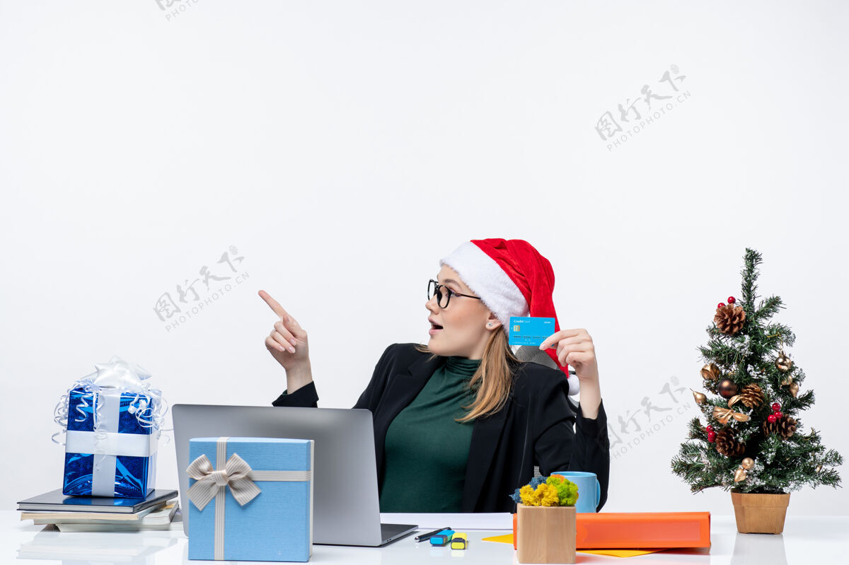 贺卡戴着圣诞老人帽戴着眼镜的漂亮女人坐在桌子旁 手里拿着圣诞礼物和银行卡在办公室里摩托板坐着眼镜
