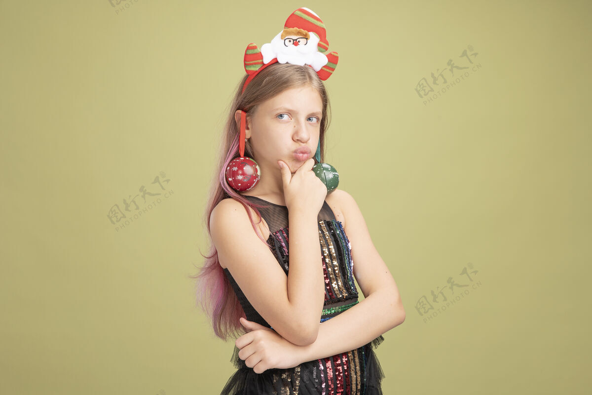头带小女孩穿着闪亮的派对礼服 头上戴着圣诞老人的头巾 耳朵上戴着圣诞球 手放在下巴上 站在绿色的墙上严肃地思考着圣诞老人圣诞节衣服