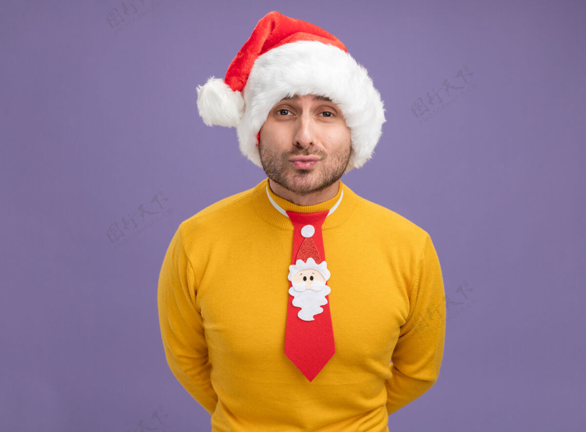 手自信的白人年轻人戴着圣诞帽 打着领带 看着摄像机 双手放在背后 做着亲吻的手势 紫色背景上 有着独立的复制空间领带亲吻看