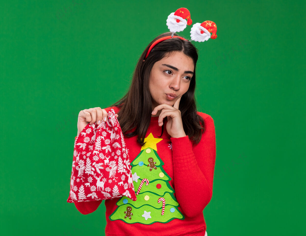抱体贴的年轻白种女孩戴着圣诞老人的头带 手指放在下巴上 拿着圣诞礼品袋 看着绿色背景上孤立的一面 还有复制空间头带年轻放