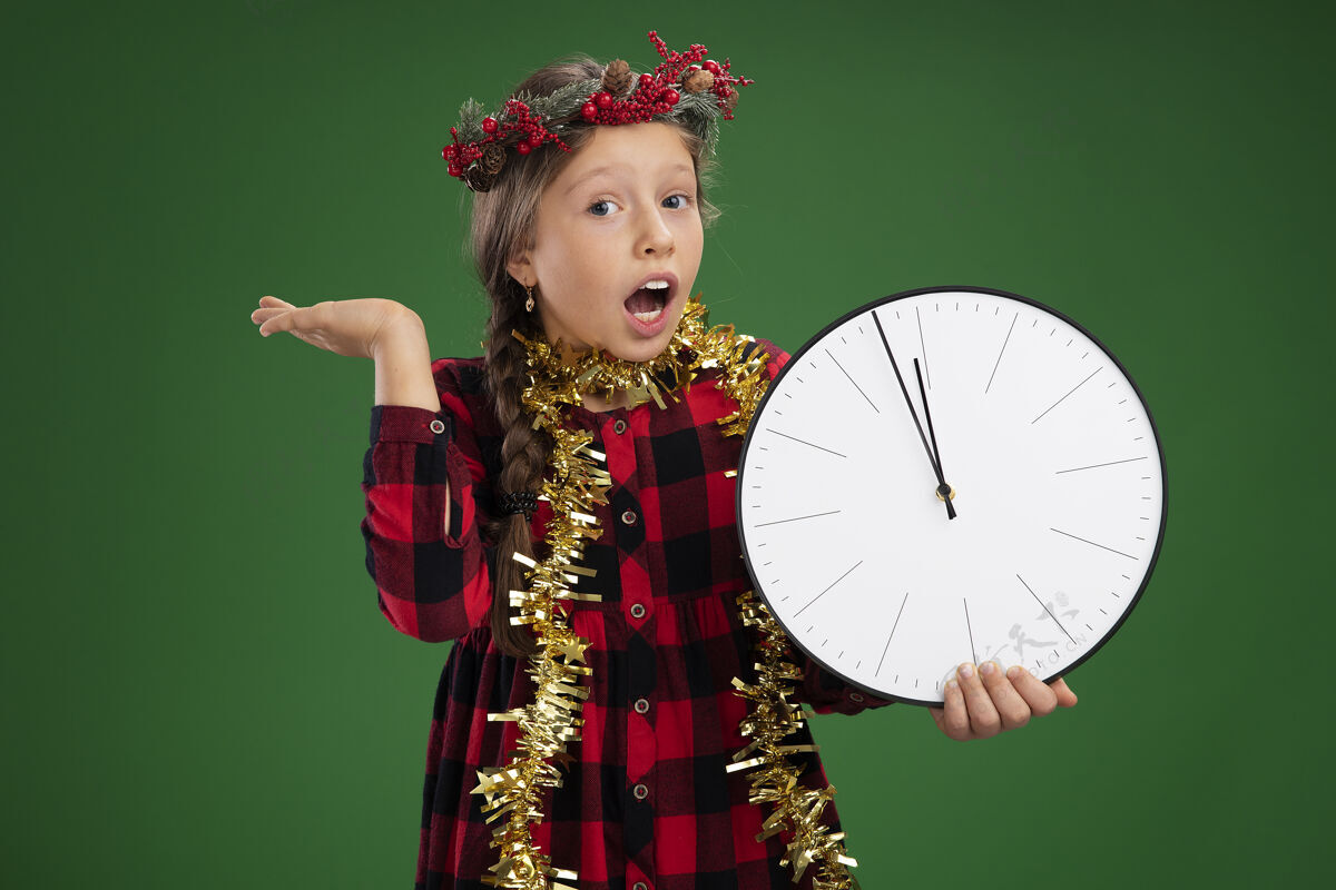 女孩情绪激动的小女孩戴着圣诞花环 身穿格子裙 脖子上戴着金属丝 手拿挂钟 看着照相机 惊讶而惊喜地举起手臂站在绿色背景上墙壁小圣诞节