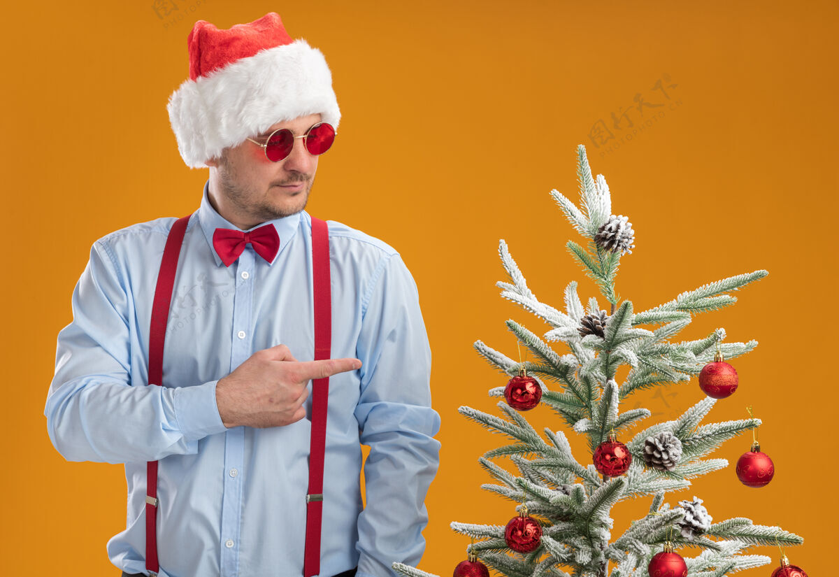 指着一个戴着圣诞帽 戴着红眼镜 系着吊带领结的年轻人站在圣诞树旁 用食指指着橙色的墙上的圣诞树年轻人吊带男人