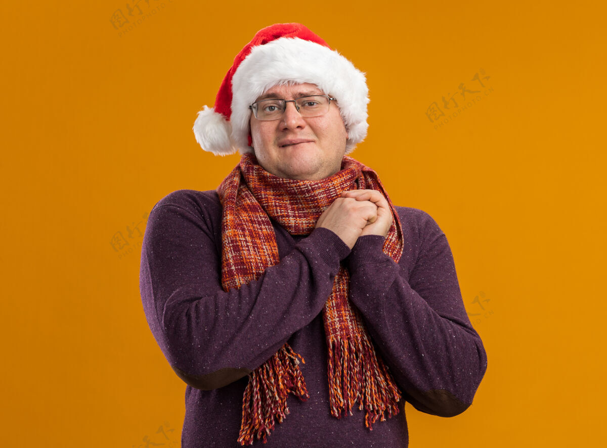 兴奋兴奋的成年男子戴着眼镜 戴着圣诞帽 脖子上围着围巾 看着镜头 手放在一起 咬着嘴唇 在橙色的背景上孤立着嘴唇一起手