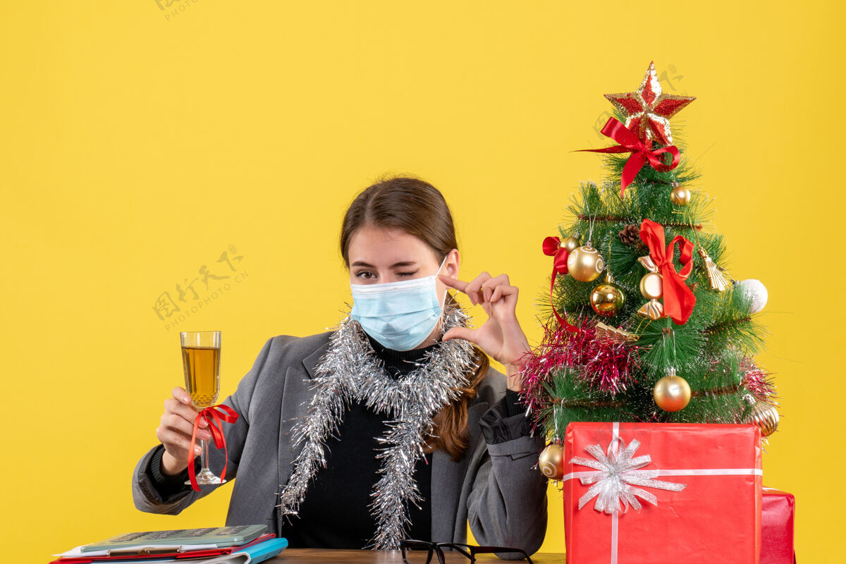 眨眼正面图：戴着医用面罩的年轻女孩坐在桌旁眨着眼睛提议敬酒 圣诞树和礼物鸡尾酒女孩人医学
