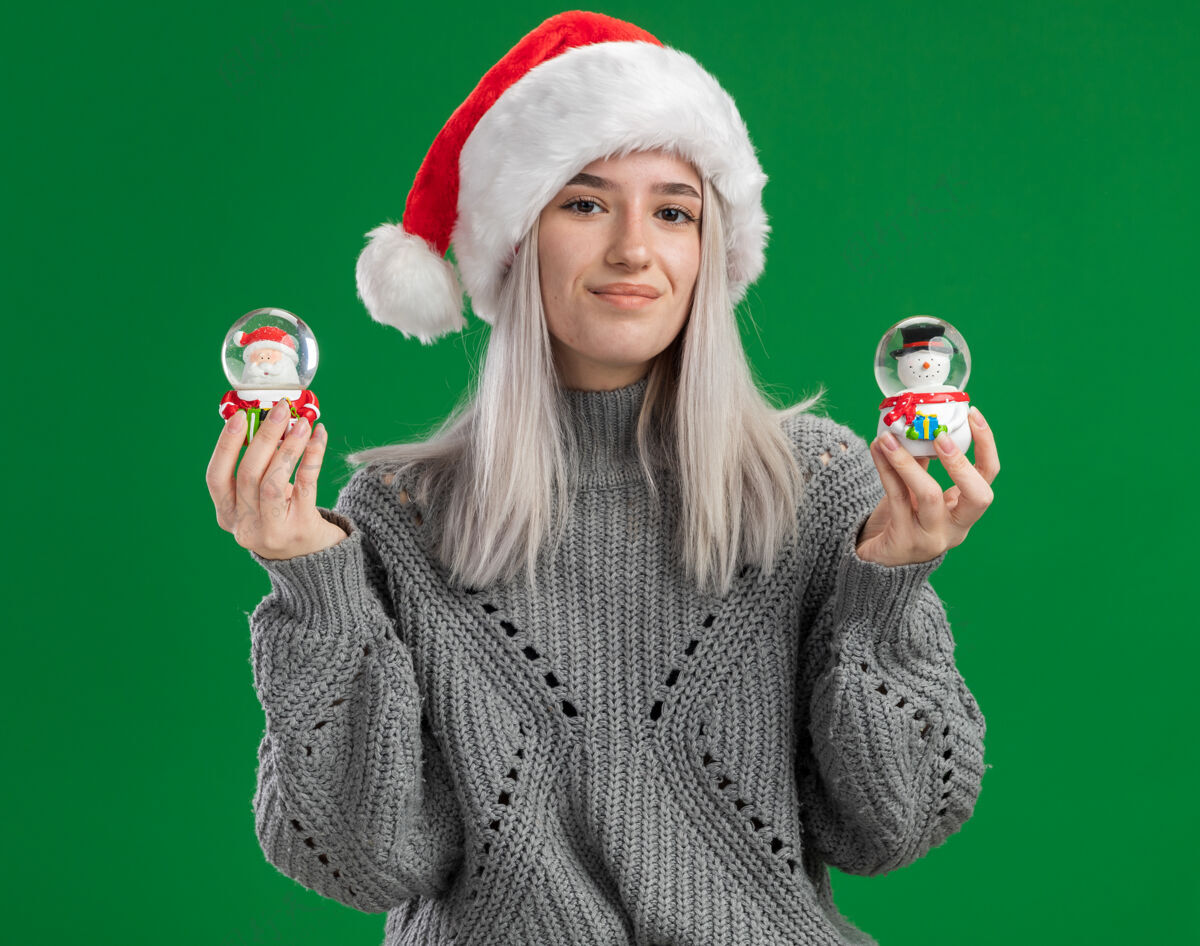 毛衣穿着冬季毛衣 戴着圣诞帽 拿着圣诞玩具雪球的快乐金发女郎站在绿色背景下 看着相机 开心地微笑着地球仪年轻帽子