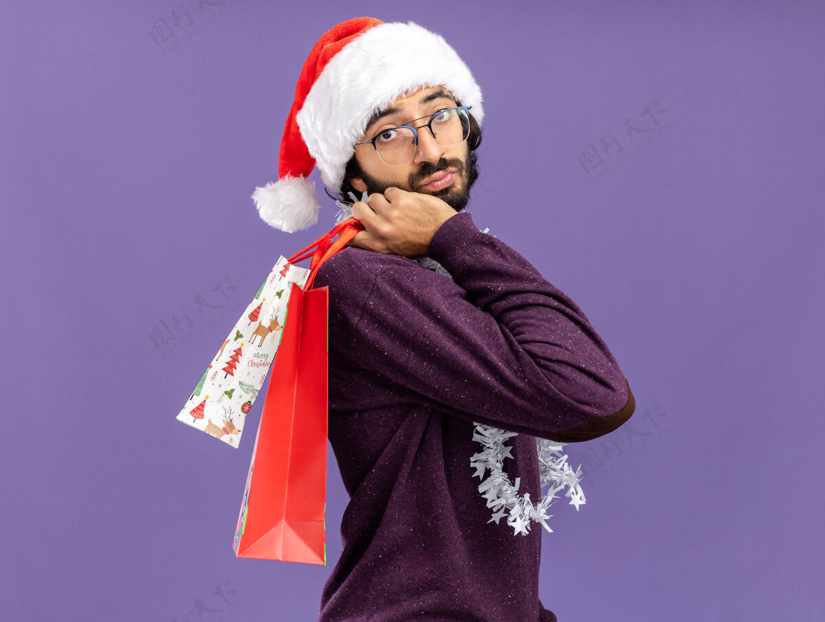礼物年轻帅哥戴着圣诞帽 脖子上戴着花环 肩上挎着礼品袋 蓝色背景 与世隔绝圣诞节小伙子肩膀