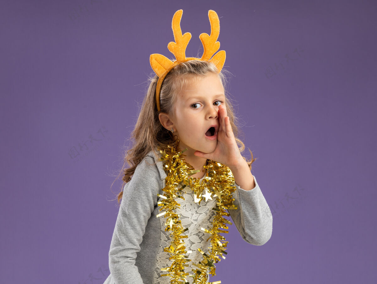 小可疑的小女孩戴着圣诞发箍 脖子上戴着花环 在蓝色背景上低语着圣诞节脖子耳语