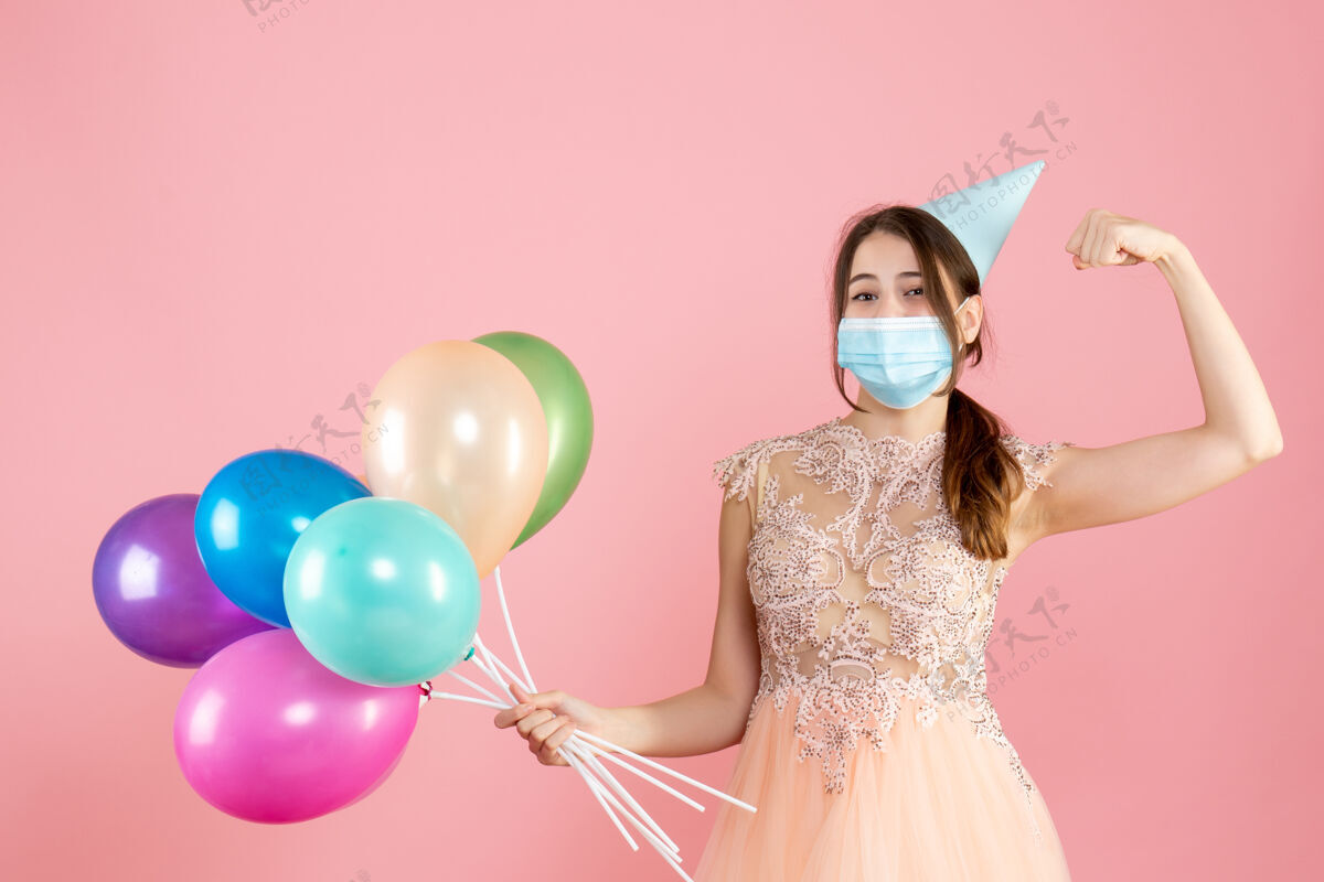 女孩戴着派对帽和医用面具的快乐女孩展示着肌肉 手里拿着粉色的彩色气球帽子气球气球
