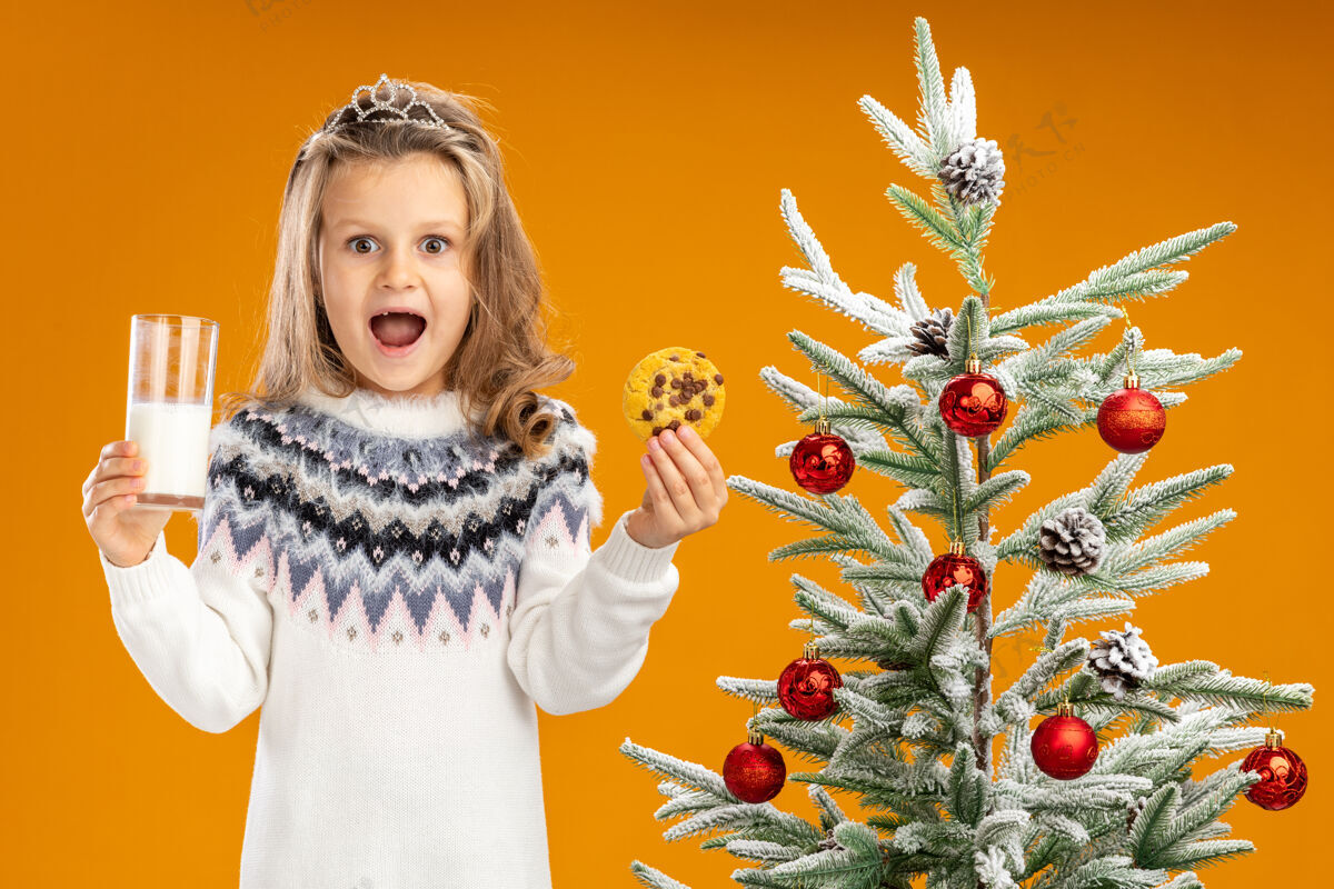 就在附近快乐的小女孩站在圣诞树旁 戴着头饰 脖子上戴着花环 手里拿着一杯牛奶 橘黄色的墙上隔着饼干快乐小戴着