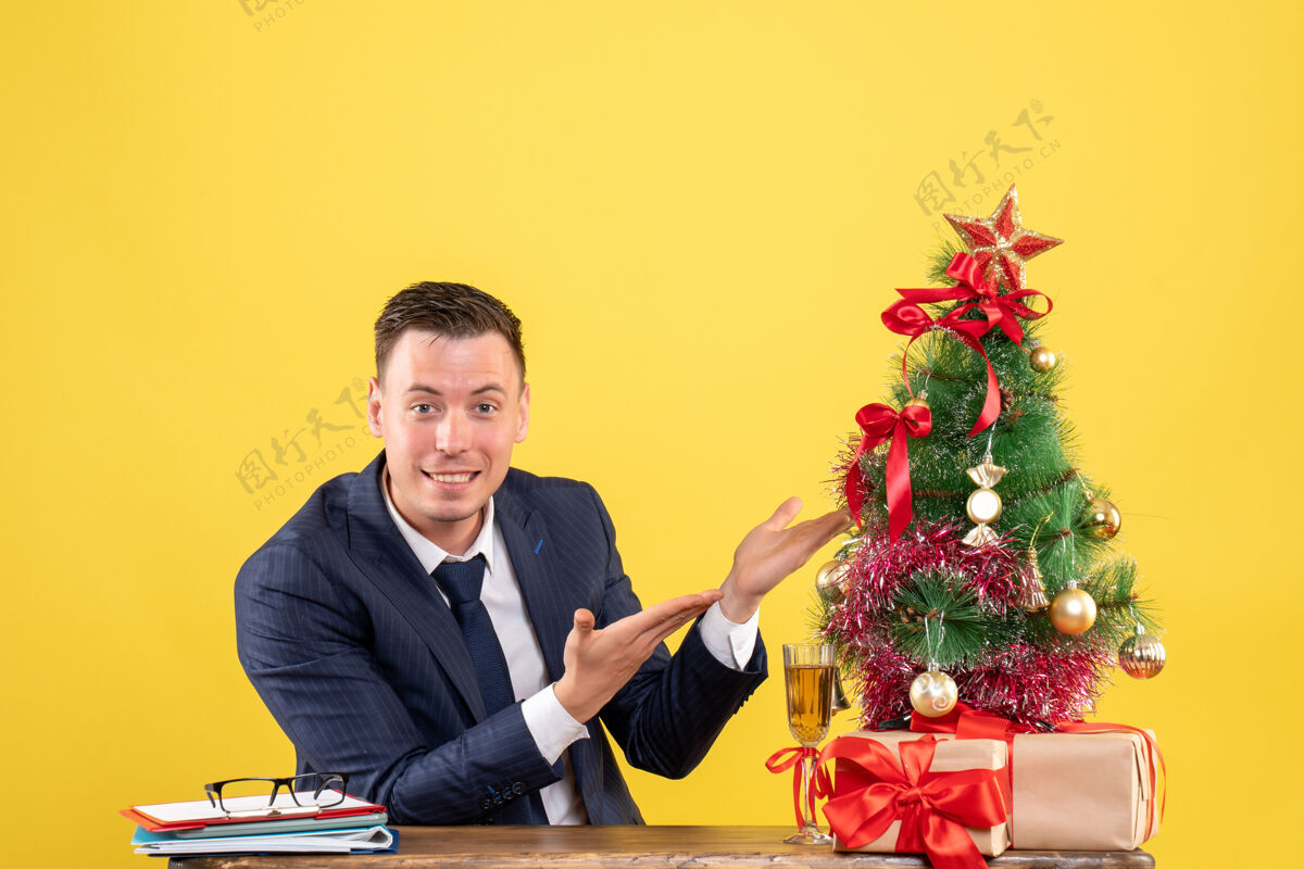 指向快乐的男人指着圣诞树坐在圣诞树旁边的桌子上 黄色的礼物桌子电脑西装