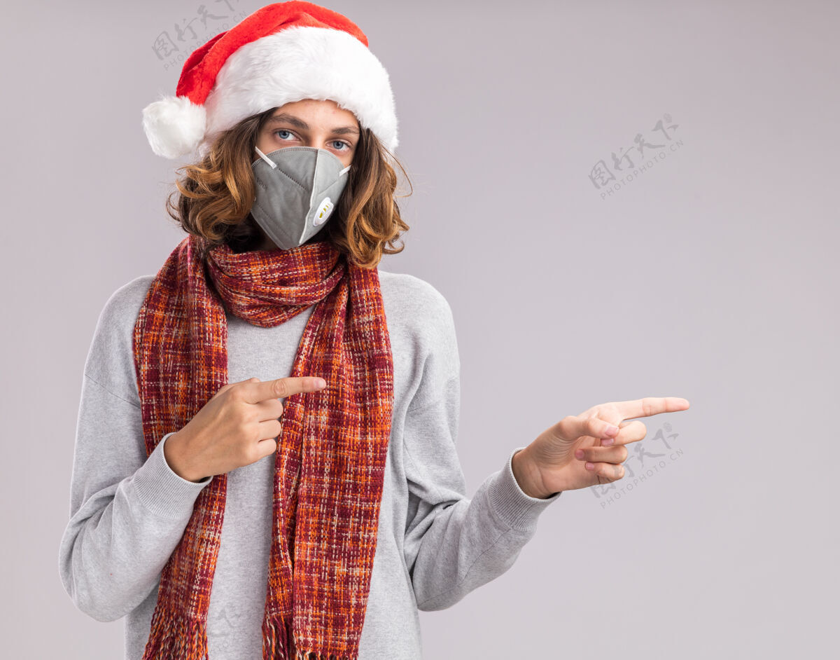 圣诞快乐年轻人戴着圣诞老人帽 戴着护面面具 脖子上围着暖和的围巾 看着摄像机 用食指指着站在白色背景上的一边站着快乐圣诞老人