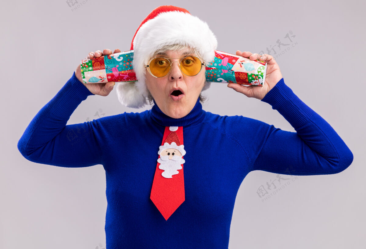 惊讶戴着太阳眼镜 戴着圣诞帽 系着圣诞领带 拿着纸杯 靠近耳朵 在白色背景上有复印空间的惊讶老妇人圣诞老人杯子耳朵