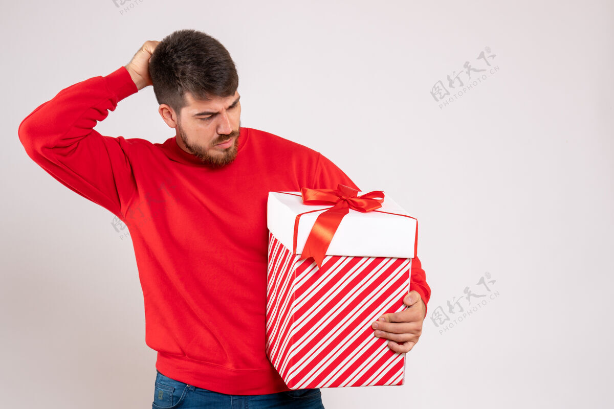 肖像身穿红衫的年轻人在白色墙上的盒子里拿着圣诞礼物前面微笑礼物