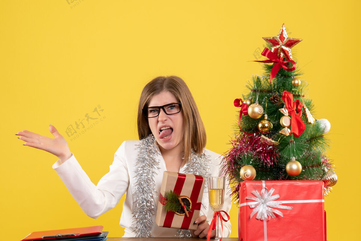 抱正面图：女医生坐在桌子前 手里拿着黄色背景的礼物 还有圣诞树和礼品盒前面圣诞节健康