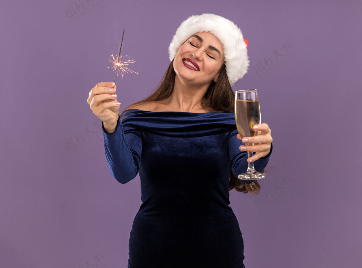圣诞节微笑着的年轻漂亮女孩 穿着蓝色连衣裙 戴着圣诞帽 手里拿着闪闪发光的东西 手里拿着一杯香槟 对着紫色背景上的镜头闪闪发光香槟相机