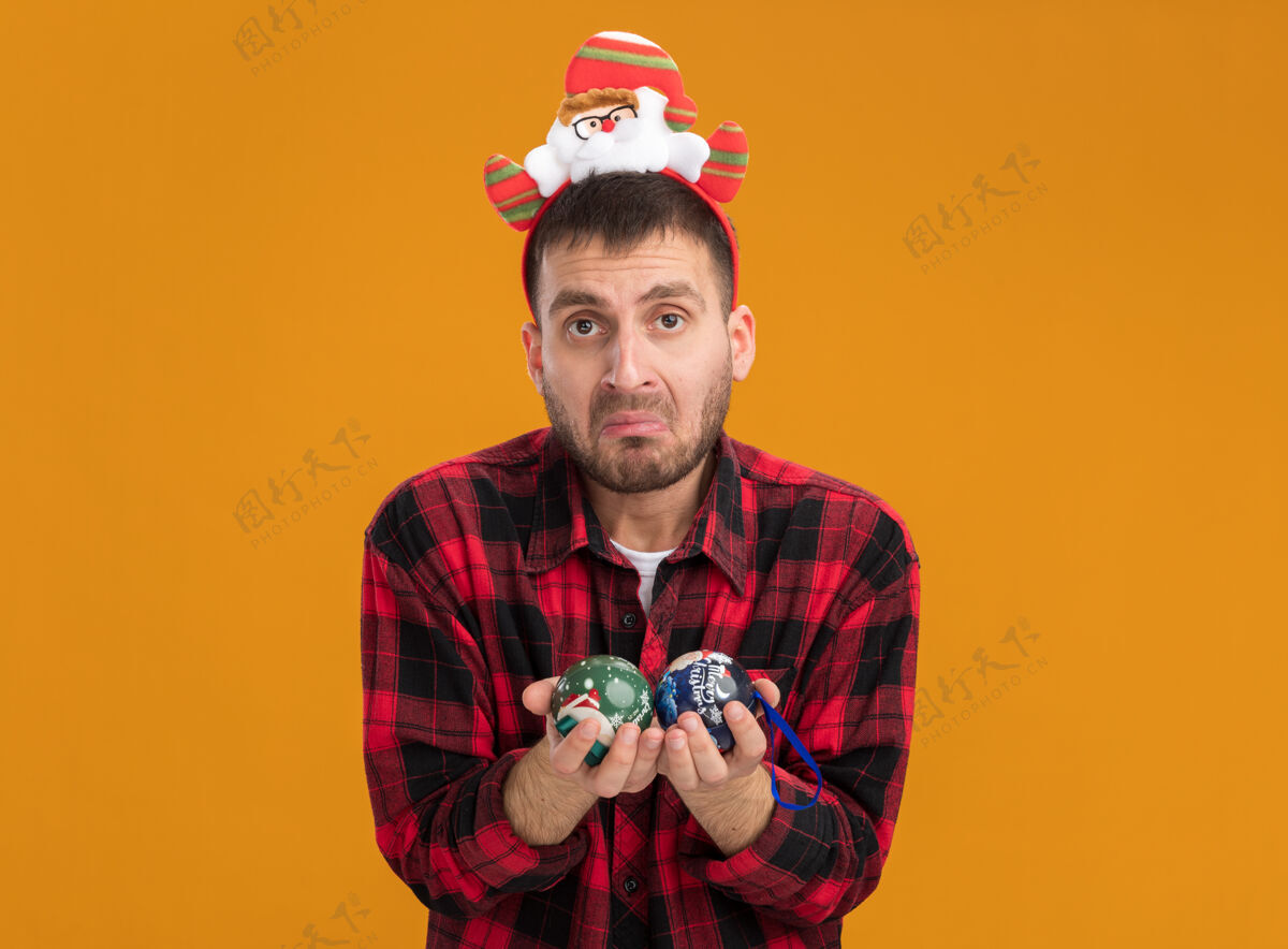 头带无知的白人年轻人戴着圣诞老人的头带 手里拿着圣诞饰品 被隔离在橙色的墙上 留着复制空间戴着圣诞老人年轻人