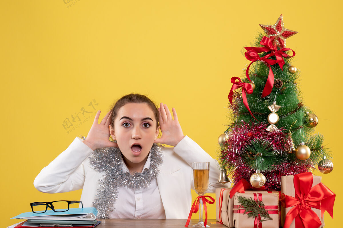 坐前视图：女医生坐在黄色背景的桌子后面 带着圣诞树和礼品盒礼物女漂亮