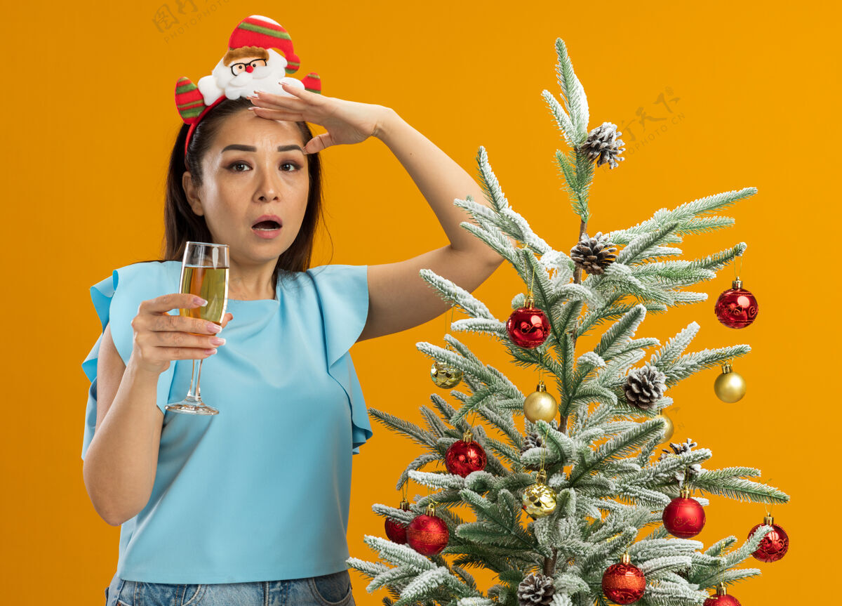 香槟身穿蓝色上衣的年轻女子头上戴着有趣的圣诞戒指 手里拿着一杯香槟 远远望去 惊讶地站在橙色背景下的圣诞树旁远头抱着