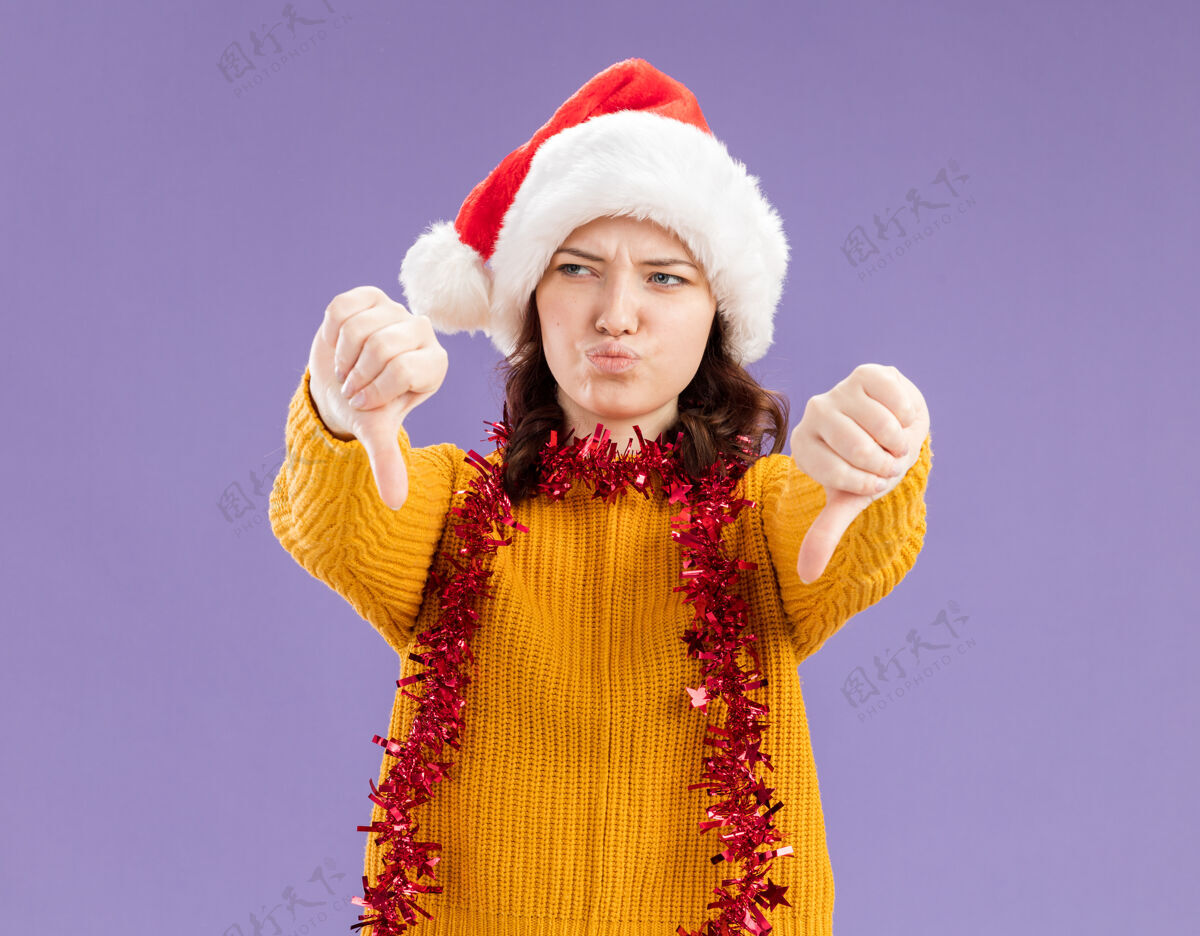 下戴着圣诞帽 脖子上戴着花环 拇指朝下 在紫色背景上孤立着 留着复制空间的未出租的年轻斯拉夫女孩年轻圣诞老人拇指