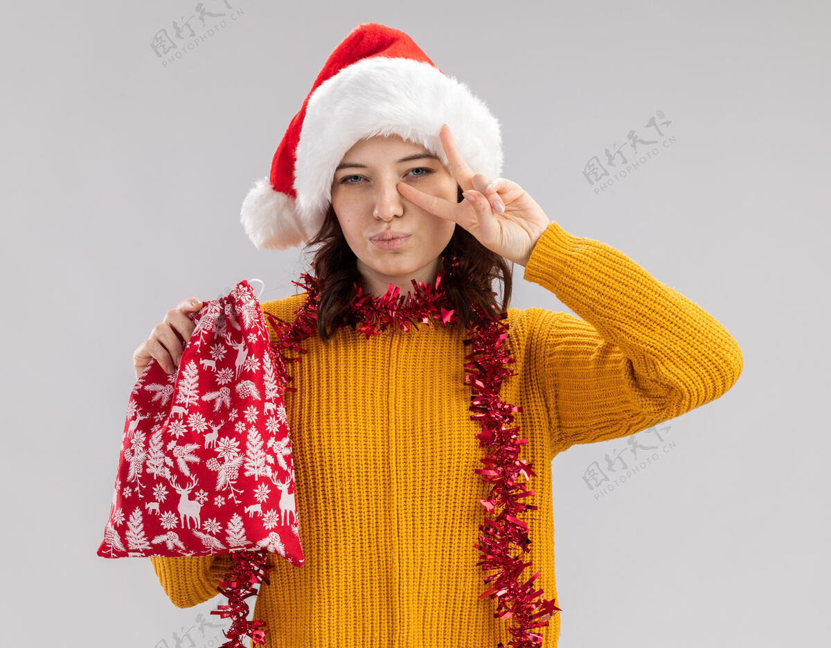 胜利自信的年轻斯拉夫女孩 戴着圣诞帽 脖子上戴着花环 手里拿着圣诞礼品袋 打着胜利的手势 白色背景上有复制空间举行周围圣诞节