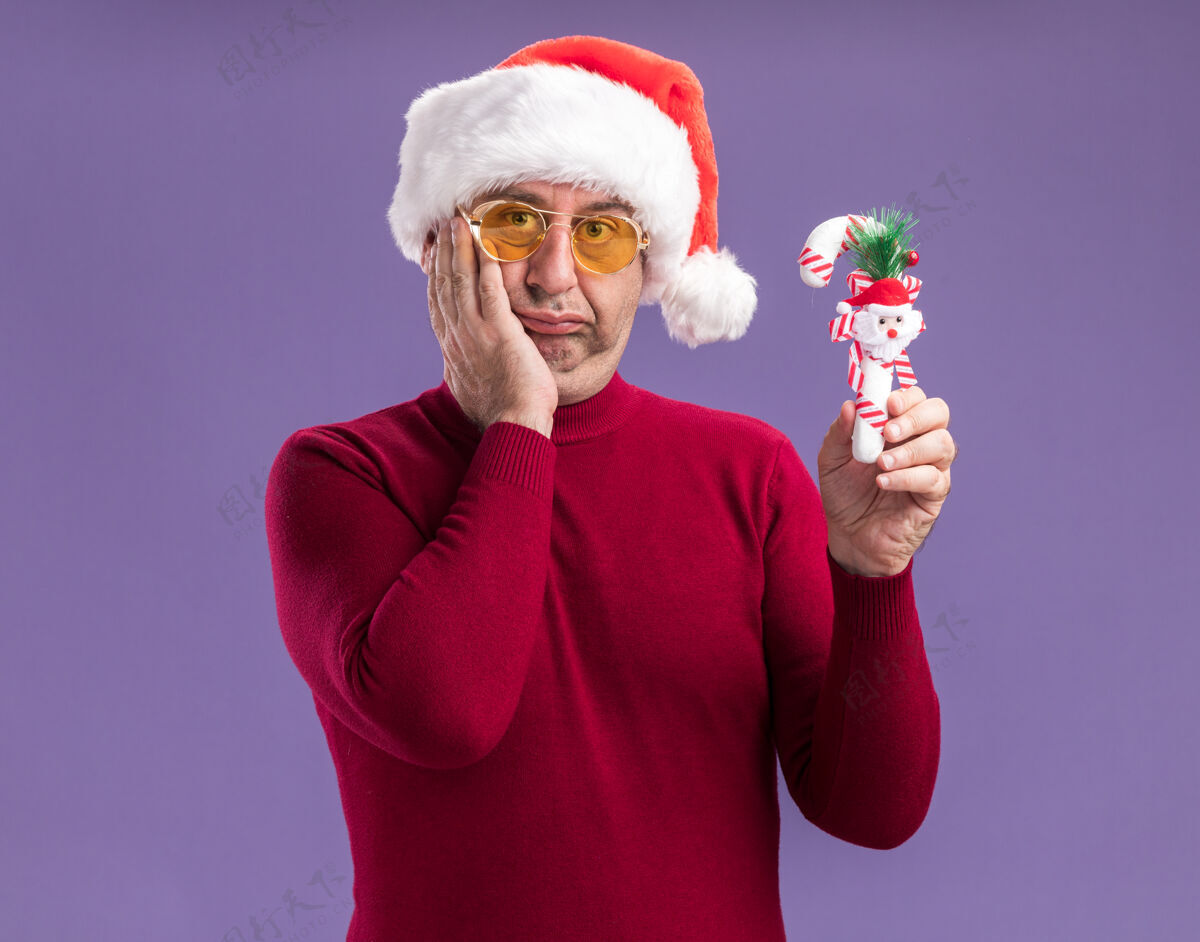 手杖中年男子戴着圣诞帽戴着黄色眼镜拿着圣诞糖果手杖困惑地站在紫色背景下看着相机抱着帽子圣诞节