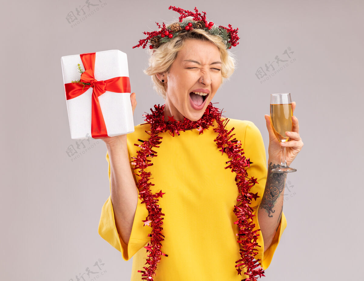 花环快乐的金发女郎戴着圣诞花环 脖子上戴着金箔花环 手里拿着一杯香槟和礼包 闭着眼睛在白色的墙上尖叫尖叫香槟金发