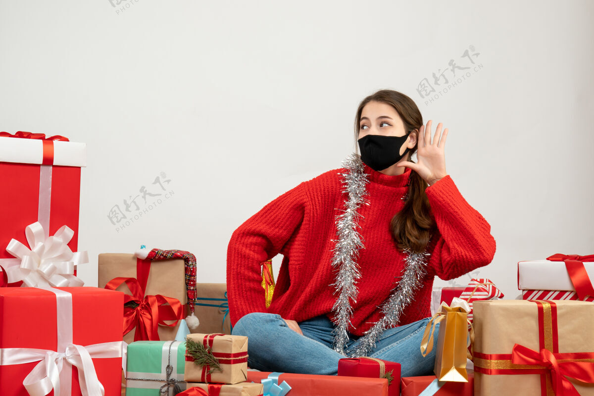 年轻女孩小女孩穿着红色毛衣 戴着黑色面具 手放在耳边 围坐在白色的圣诞礼物上红色成人礼物