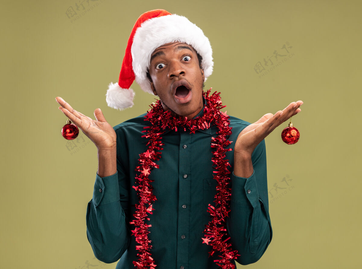 举行非洲裔美国人戴着圣诞帽 戴着花环 手里拿着圣诞球 站在绿色的背景上 惊讶地看着镜头圣诞老人美国圣诞节