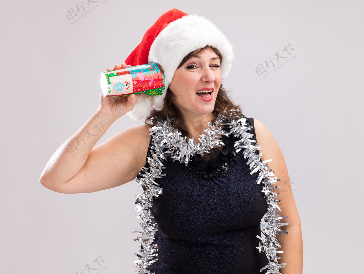 举行好奇的中年妇女戴着圣诞帽 脖子上戴着金属丝花环 手里拿着塑料圣诞杯 耳边听着秘密 看着相机 在白色背景上眨眼杯子中年脖子