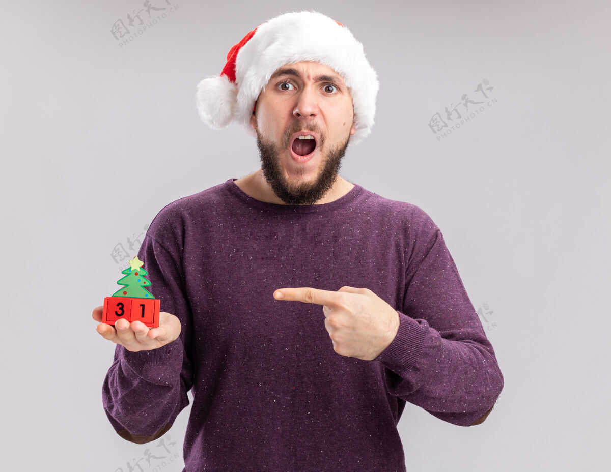 惊讶身穿紫色毛衣 戴圣诞帽的年轻人站在白色背景上 用食指指着方块上的新年日期 既惊讶又惊讶站着圣诞老人日期