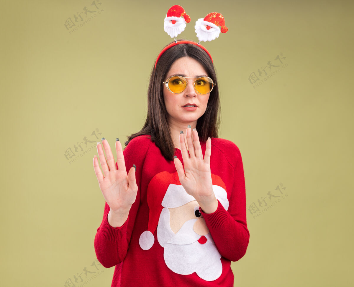 圣诞老人年轻漂亮的白人女孩 穿着圣诞老人的毛衣 戴着帽子 戴着眼镜 在橄榄绿的背景上做着一个孤立的手势 还有复印空间橄榄头带拒绝