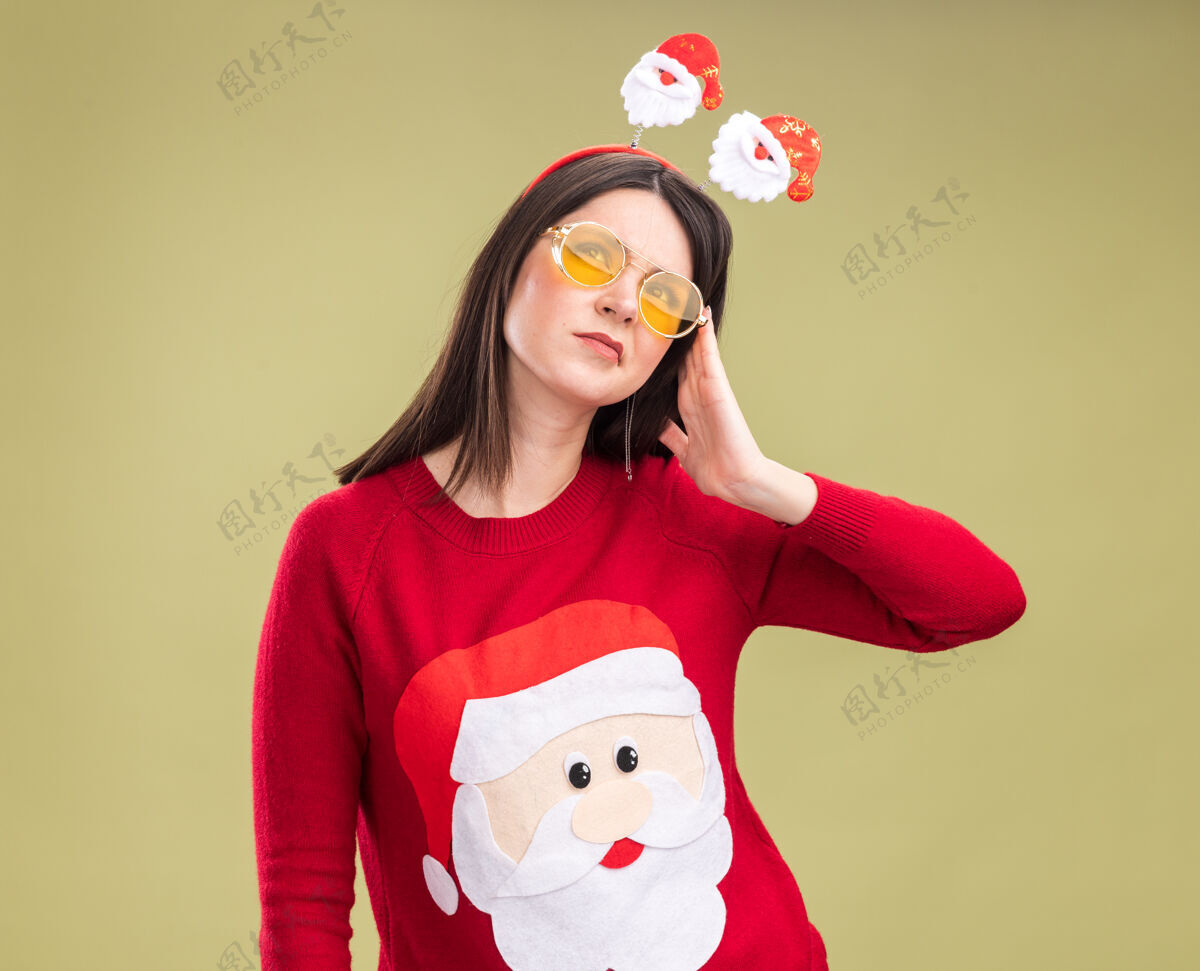 触摸困惑的年轻漂亮的白人女孩穿着圣诞老人的毛衣和头带 戴着眼镜 在橄榄绿的背景下孤立地抬头看头橄榄困惑