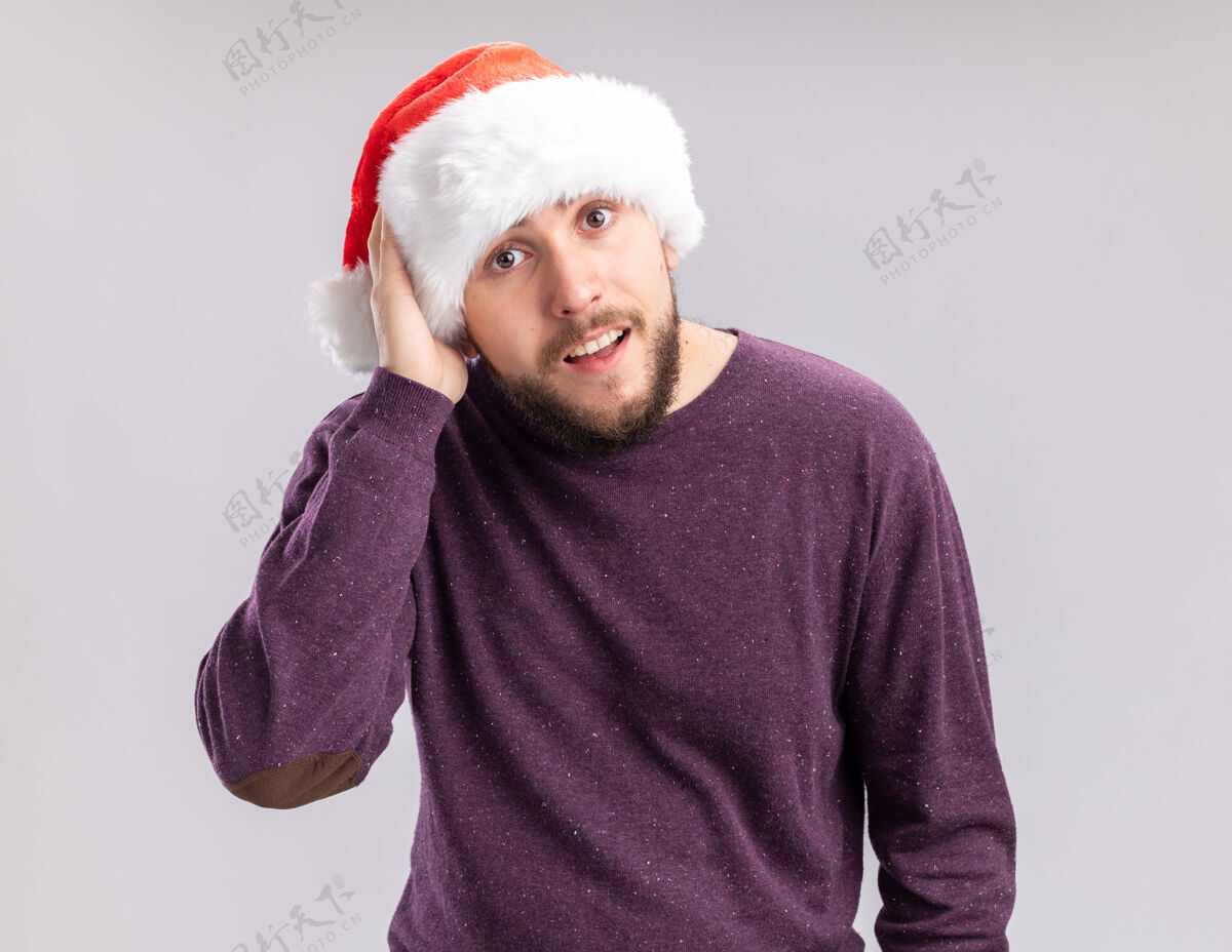 年轻一个穿着紫色毛衣 戴着圣诞帽的年轻人站在一边看着相机 困惑地用手在他的头上画着白色的背景站着手圣诞老人
