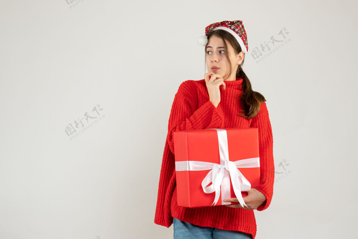 东西戴着圣诞帽的窥探女孩拿着礼物看着站在白色地板上的东西人圣诞节站着