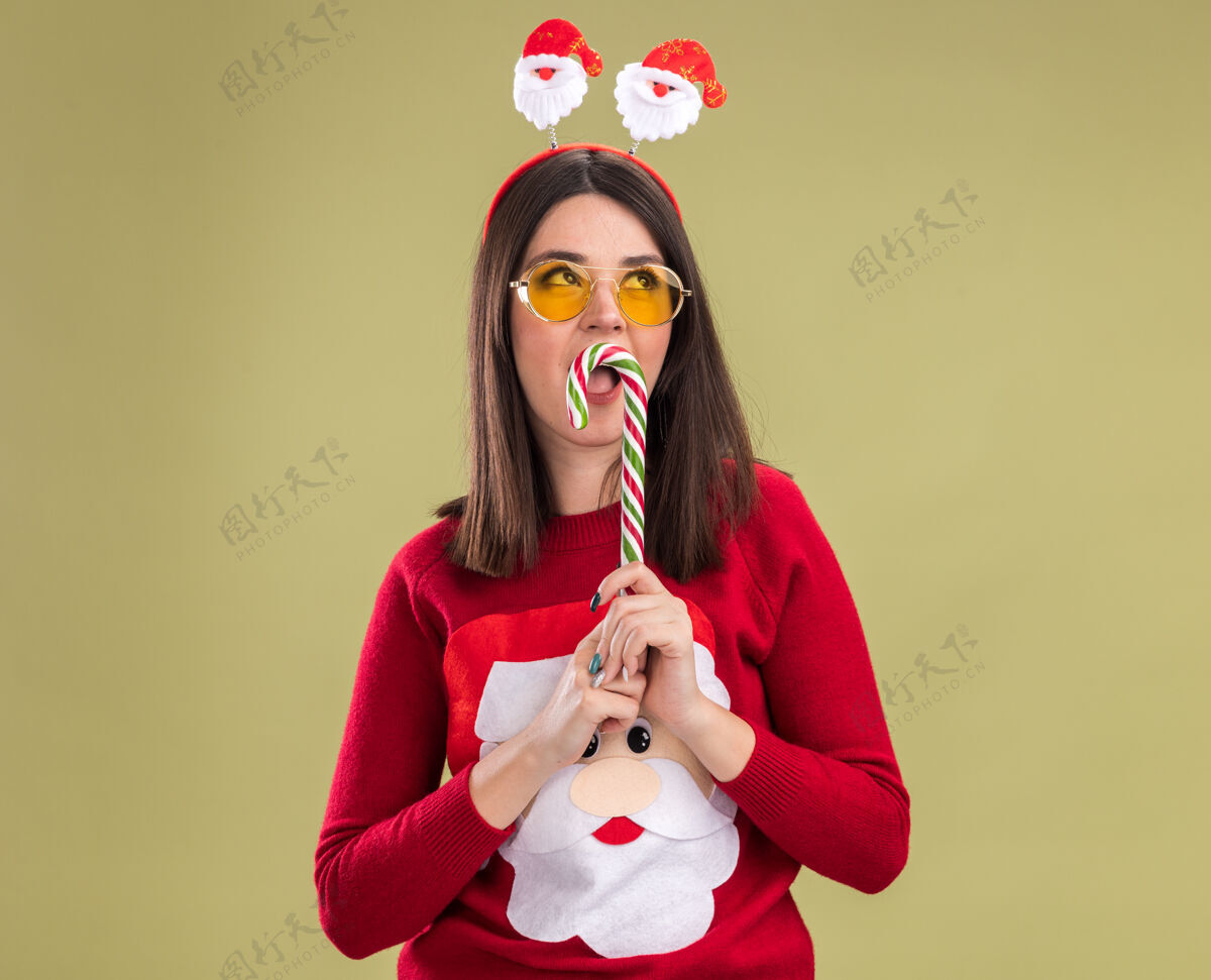 快乐年轻漂亮的白人女孩 穿着圣诞老人毛衣 头戴眼镜 嘴里拿着传统的圣诞糖果手杖看嘴绿色