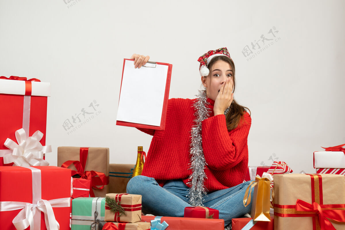 惊喜惊讶可爱的女孩拿着圣诞帽拿着文件坐在白色的礼物周围成人家可爱