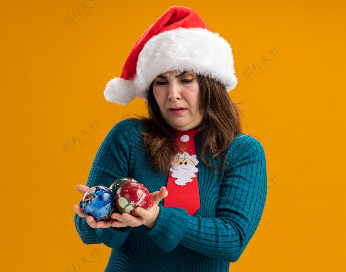 圣诞老人恼怒的成年白人妇女拿着圣诞帽和圣诞领带 看着橙色背景上的玻璃球饰品 还有复制空间领带橙色球