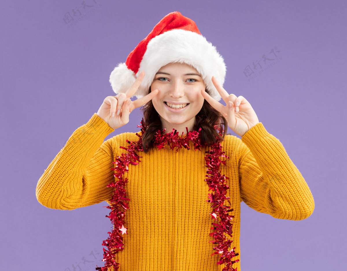 年轻微笑着的年轻斯拉夫女孩 戴着圣诞帽 脖子上戴着花环 在紫色的背景上用复制的空间表示胜利的标志手势花环斯拉夫