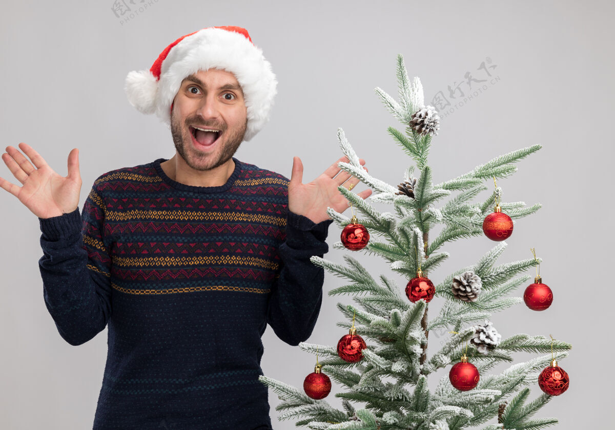 空着令人印象深刻的年轻白种人戴着圣诞帽站在圣诞树旁 空手孤立在白色的墙上展示印象深刻圣诞节