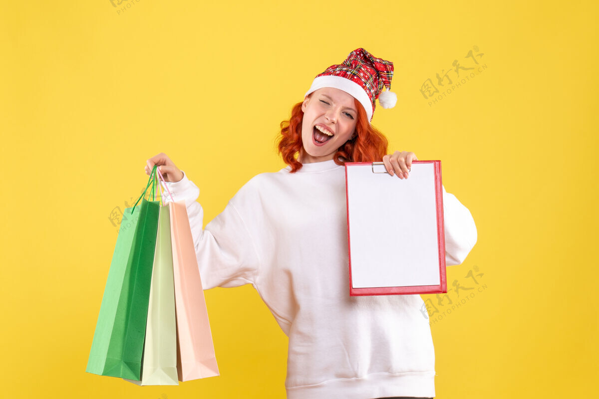 抱着黄色墙壁上拿着购物包的年轻女子的正视图节日微笑包装